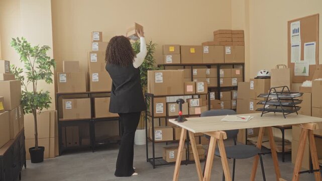 Woman tossing a cardboard box overhead while reaching up at a shipping table in a building; small business cheer.