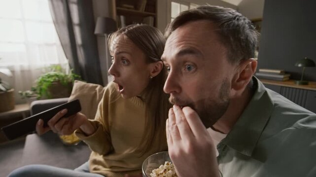 Medium close-up of young Caucasian couple sitting on sofa, watching horror flick on TV, staring at screen during moment of suspense then jumping up with sudden fear