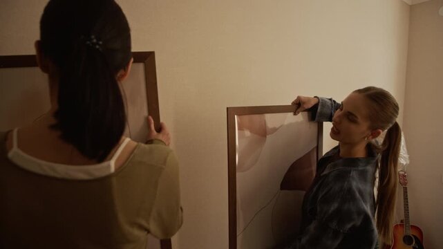 Back view medium shot of two young women trying abstract pictures on wall during moving day while two diverse men talking by window among cardboard boxes and packed furniture in new apartment