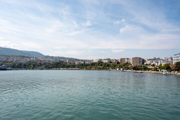 Fototapeta premium Scenic view of a Mediterranean coastal town with multi-story buildings - boats anchored in calm turquoise waters - and mountainous backdrop under partly cloudy skies