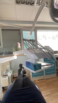 Legs in dental chair under overhead lamp with tray and instruments visible, window and plant in background, blue bib and clean clinical surfaces