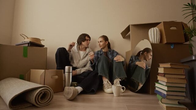 Eye level wide shot of young Caucasian couple sitting on floor among cardboard boxes and chatting in bright new apartment on moving day