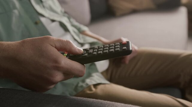 Side close-up of hand of anonymous male viewer pressing buttons to operate remote control and navigate content menu, choosing movie to watch on cable or smart TV during leisure time