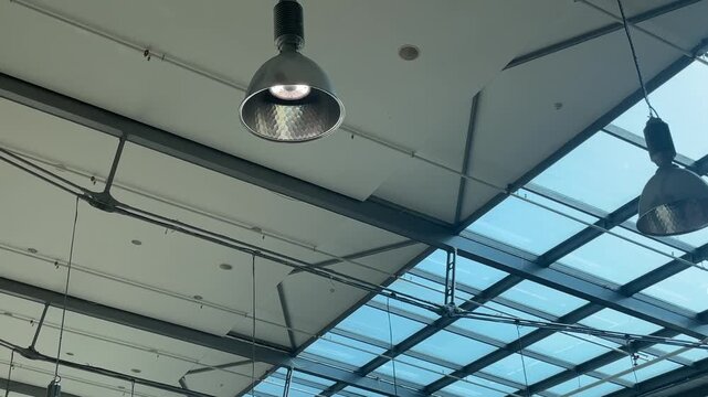 Industrial pendant lamp under skylight glass, closeup of metal fixture hanging from ceiling grid. Metal cord, reflector interior, and bluetinted glass