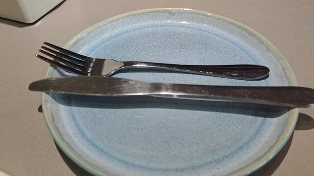 Stainless steel fork and knife resting in parallel on a blue ceramic plate. Table etiquette indicating the end of the meal at a restaurant