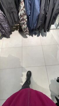 Shopping aisle view with clothing racks and tiled floor, pink jacket hem and boots visible from downward perspective, shopper browsing among coats