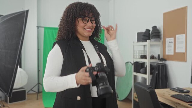 Woman photographer smiling adjusts glasses while holding dslr camera and strap in studio with green screen, softbox lights and shelving gear; creative confidence.