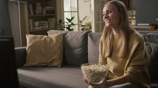 Medium shot of cheerful blonde Caucasian woman watching comedy or sitcom on cable TV at home, rolling with laughter on couch at humor and jokes, eating popcorn from bowl