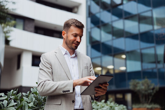 Successful businessman in a formal suit using a digital tablet while standing in front of a modern glass office building in the city