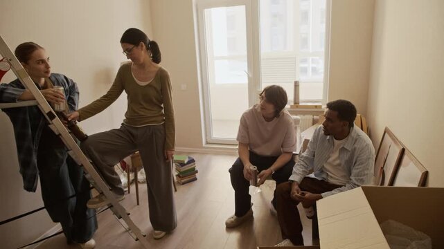 Full shot of four diverse friends standing and chatting in bright living room among cardboard boxes, packed furniture and ladder during moving day, wide interior shot showing relocation and home setup