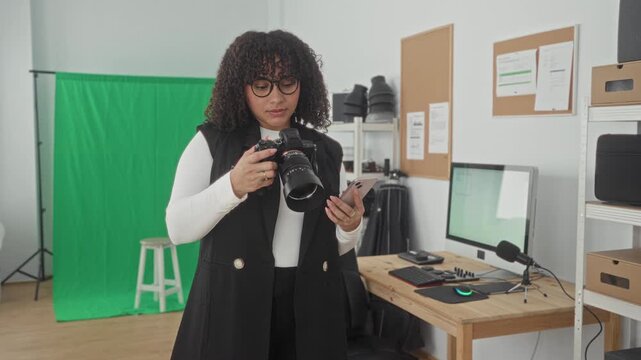 Woman holding a dslr camera and checking a smartphone in a studio workspace beside a computer desk and green screen; creative focus.