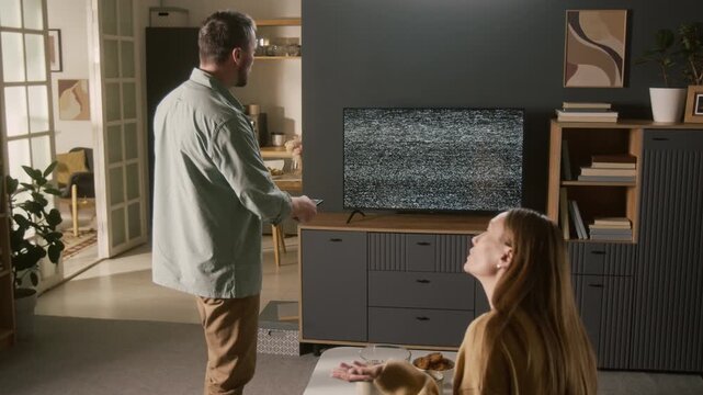 Medium full shot of frustrated young Caucasian couple arguing over broken TV displaying static interference noise on screen, husband and wife blaming each other for breaking device