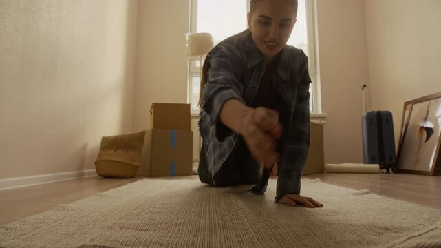 Eye level shot of young Caucasian woman unrolling rug on wooden floor among cardboard boxes while settling into bright new apartment on moving day
