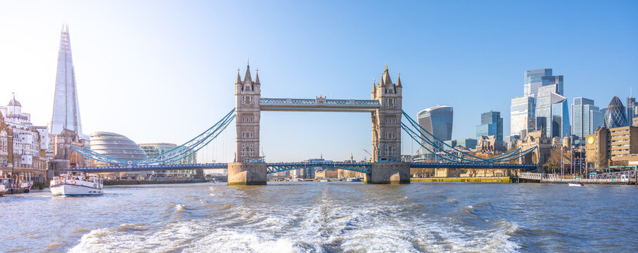 A ship moves on the river, showing Tower Bridge in London. Tall buildings fill the background under a clear sky. Water pushes away from the ship as it travels.