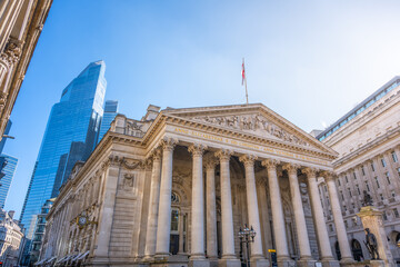 Naklejka premium The Royal Exchange in London stands among modern structures on a clear day. People walk nearby, and the British flag flies high above the historic site.