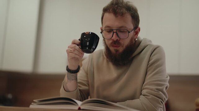 Man With Glasses Reads a Book While Sipping Coffee at Home, Enjoying Quiet Morning