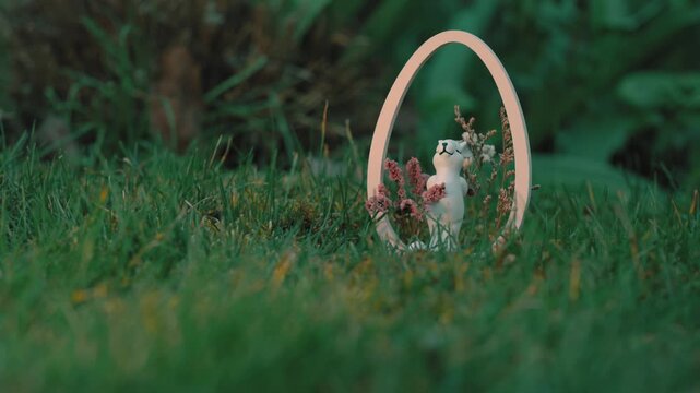 A cinematic shot of a white ceramic Easter bunny figurine nestled in a wooden oval frame on lush green grass. The scene exudes a peaceful, whimsical spring atmosphere in a garden setting.