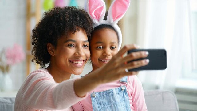 Mother and daughter taking selfie with smartphone, child wearing bunny ears headband, easter holiday at home, family bonding indoors, pastel decor background, cheerful spring celebration