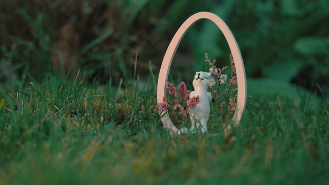 A cinematic shot of a white ceramic Easter bunny figurine nestled in a wooden oval frame on lush green grass. The scene exudes a peaceful, whimsical spring atmosphere in a garden setting.