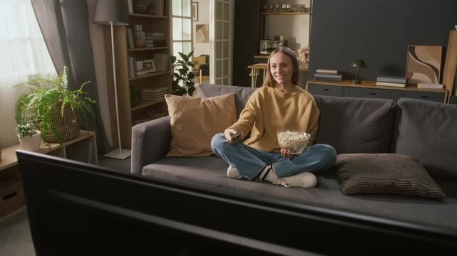 Full shot of cheerful young Caucasian woman sitting on couch with crossed legs, holding bowl of popcorn, switching TV channels with remote control, finding favorite comedy show or sitcom and watching