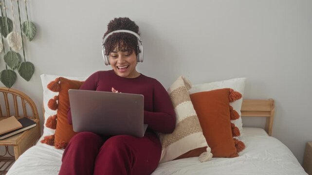 Woman typing on laptop with headphones, smiling and giving thumbs up on a bed with cushions, pillows and wall plants in studio; relaxed focus.