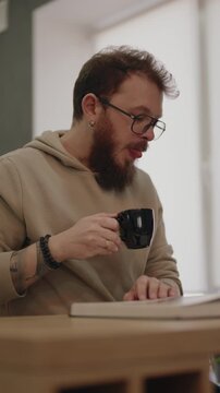 Bearded Man With Glasses Enjoys His Morning Coffee While Reading a Book at Home