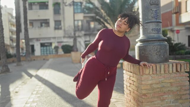 Woman in burgundy tracksuit stretching one leg against a brick pillar while holding a lamppost and balancing on the sidewalk of a sunny street; calm focus.