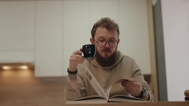 Bearded Man in Glasses Enjoys Quiet Time Reading a Book and Sipping Coffee at Home.