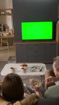 Vertical rear shot of young Caucasian father, daughter and mother watching interesting movie or sitcom on online TV streaming service with green chromakey screen, while eating popcorn