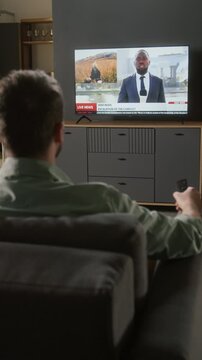 Vertical rear shot of man watching TV, switching channels to breaking news report on recent military conflict, Black male reporter speaking on geopolitics, soldiers firing weapons and storming ruins