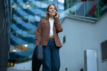 Fototapeta premium Smiling businesswoman talking on phone walking in urban environment