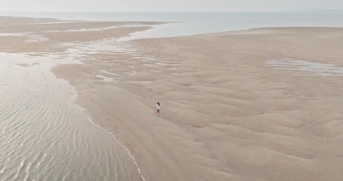 Explore Koh Lanta, Thailand: Woman walking barefoot across vast low tide sandbar sand beach, embracing solitude and open tropical coastline with serene sea, sky and rippled sandy texture. Aerial view