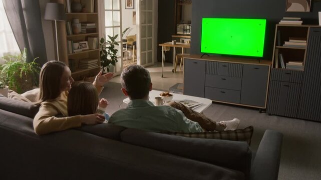 Full rear shot of excited young Caucasian family sitting on couch at home, watching fun movie, comedy or cartoon on smart TV streaming service, pointing at chromakey green screen