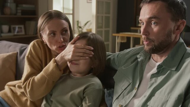 Medium shot of young Caucasian parents and daughter watching movie together on TV, shocked by explicit inappropriate scene, mother covering child eyes while arguing with father