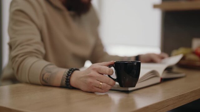 Man Reading a Book While Holding a Coffee Cup on a Wooden Table