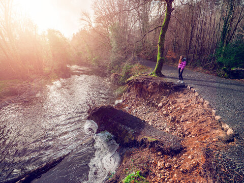 Young woman is looking at a scale of damaged river bank of a forest park after a strong hurricane or storm. Big chunk of earth and walking path was damaged and fallen into a fast creek. Warm sunlight.