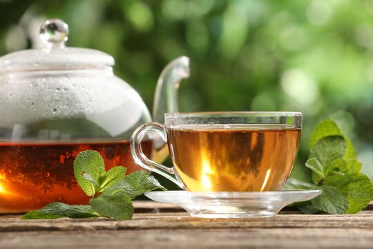 Aromatic tea in glass cup, teapot and mint on wooden table outdoors, closeup