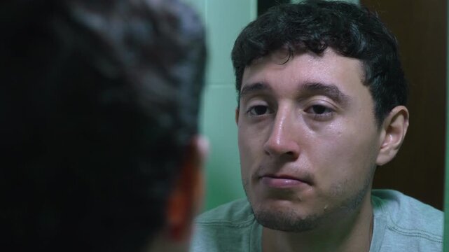 Hombre joven mir&aacute;ndose al espejo con rostro h&uacute;medo en ba&ntilde;o lavandose la cara