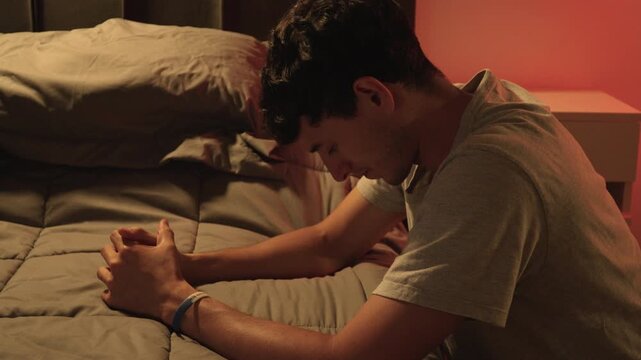 Hombre joven orando junto a la cama en habitaci&oacute;n