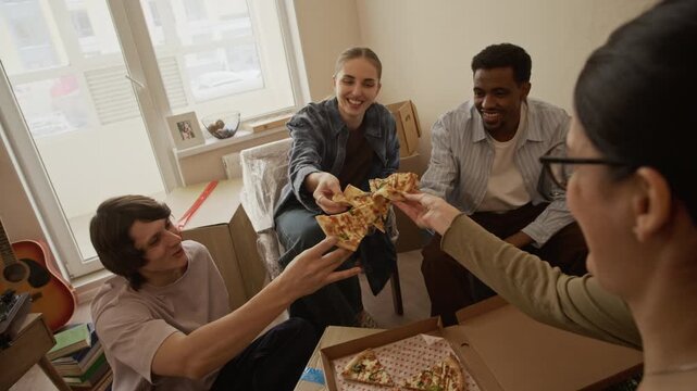 Dolly out shot of four diverse friends relaxing on floor and toasting with takeaway pizza slices, sitting among cardboard boxes in bright apartment on moving day