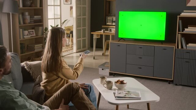 Dolly-in shot of young Caucasian woman and man choosing movie to watch on smart TV with green chromakey screen, wife switching channels with remote control or navigating menu on streaming service