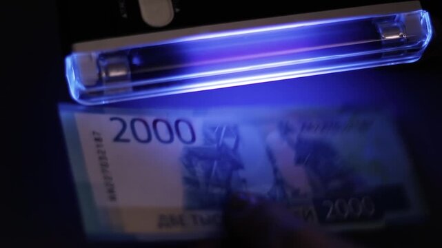 A man checks the authenticity of a two-thousand-ruble bill under an ultraviolet light. Video