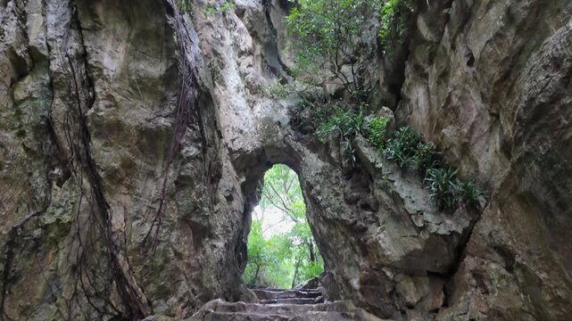 A stone staircase leads through a natural archway in the Marble Mountains, a popular tourist destination in Da Nang, Vietnam. Lush greenery surrounds the opening.