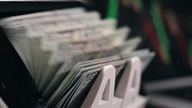 Cash counting machine processes stacks of hundred-dollar bills, displaying the continuous flow of currency in a financial setting with digital graphs in the background