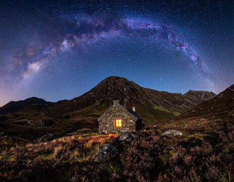 Traditional stone bothy illuminated at night under the Milky Way in the Scottish Highlands. Sharp mountain silhouette, heather texture, and starry sky. Concept for sanctuary and vastness.