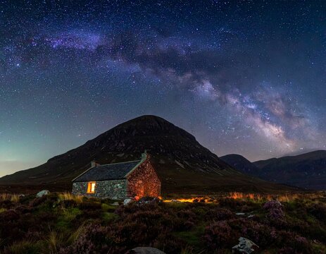 Traditional stone bothy illuminated at night under the Milky Way in the Scottish Highlands. Sharp mountain silhouette, heather texture, and starry sky. Concept for sanctuary and vastness.