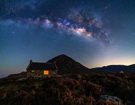 Traditional stone bothy illuminated at night under the Milky Way in the Scottish Highlands. Sharp mountain silhouette, heather texture, and starry sky. Concept for sanctuary and vastness.