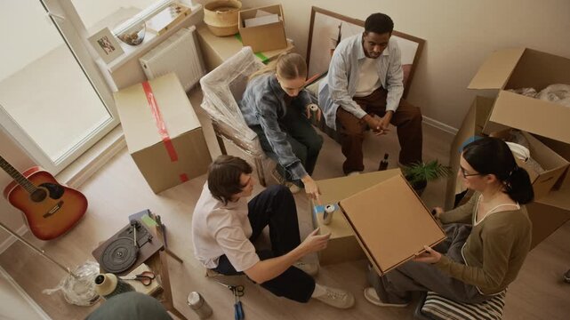 High angle shot of four multiethnic friends sitting on floor enjoying takeaway pizza and conversation among cardboard boxes in bright apartment on moving day