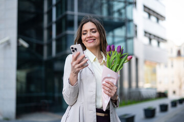 Fototapeta premium Smiling businesswoman holding a bouquet of tulips while looking at her smartphone outdoors