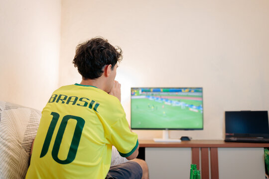 Torcedor brasileiro assistindo a uma partida internacional de futebol em casa, visto de costas com a camisa da sele&ccedil;&atilde;o. Jovem focado na TV em momento decisivo, transmitindo tens&atilde;o e expectativa.